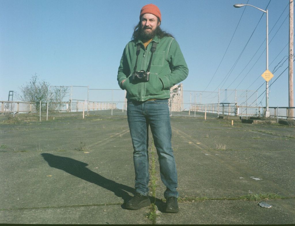 Adam stands on an empty paved lot wearing an orange knit cap and green